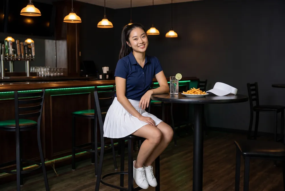 Generated picture of the same character on a tall stool at a high table in a dark bar and grille, a glass of sparkling water and a plate of fries beside her white visor.